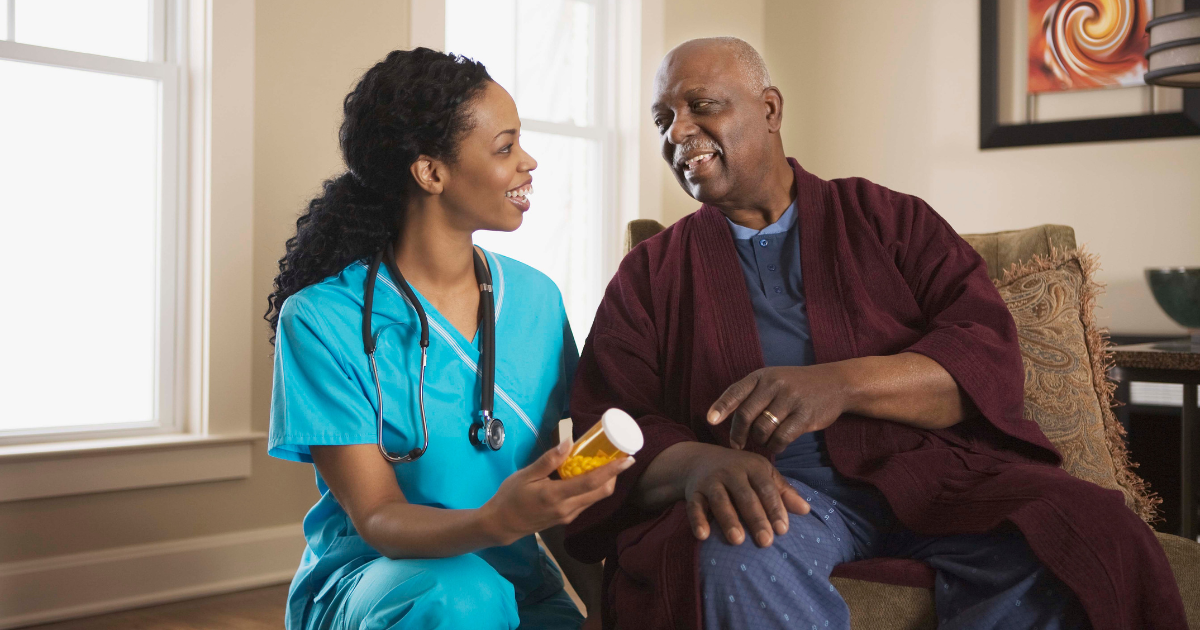 A home care nursing assistant kneels next to an elderly man holding a pill bottle