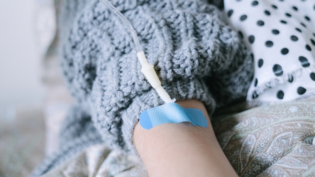 A close-up of a person's arm receiving intravenous therapy, with a medical IV line inserted and secured using a blue adhesive patch. The individual is wearing a cozy gray knitted sweater and resting on patterned bedding. This image represents intravenous therapy in Temple, TX, emphasizing comfort and care during treatment.