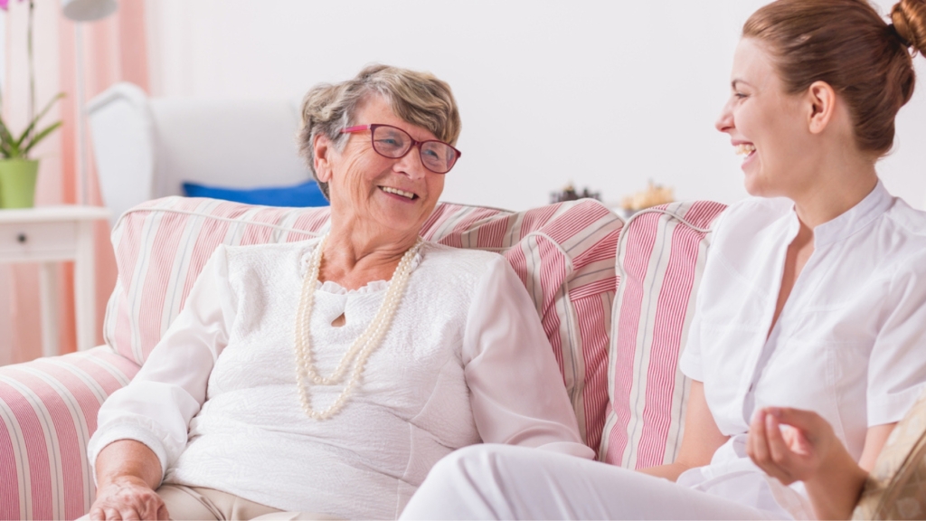 A female caregiver smiles warmly while seated on a couch beside a senior woman, assisting her with speech therapy. The caregiver holds a set of flashcards as they engage in a supportive session, reflecting compassionate speech therapy service in Temple, TX.