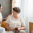 Smiling caregiver with her hand on an older woman’s shoulder as they share tea on the couch, showing home care vs assisted living