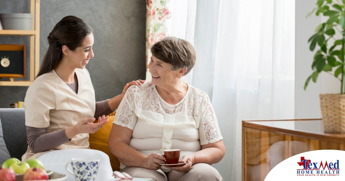 Smiling caregiver with her hand on an older woman’s shoulder as they share tea on the couch, showing home care vs assisted living